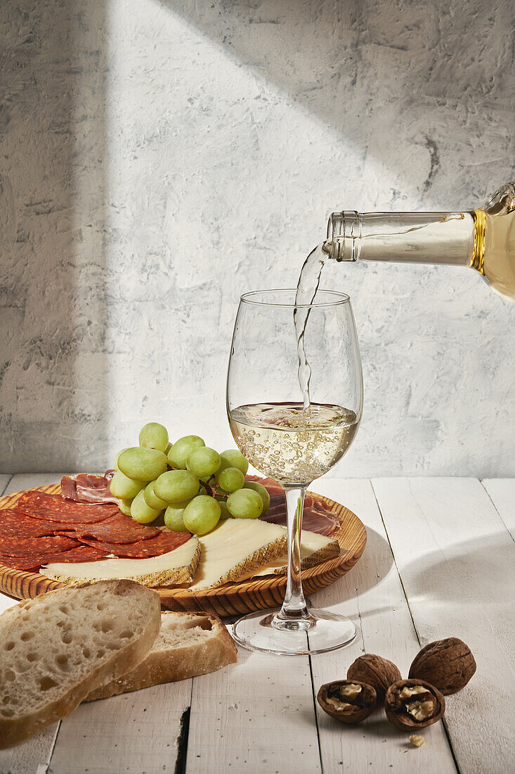 Composition of flavoring white wine pouring into wineglass and served on table near meat platter with grapes
