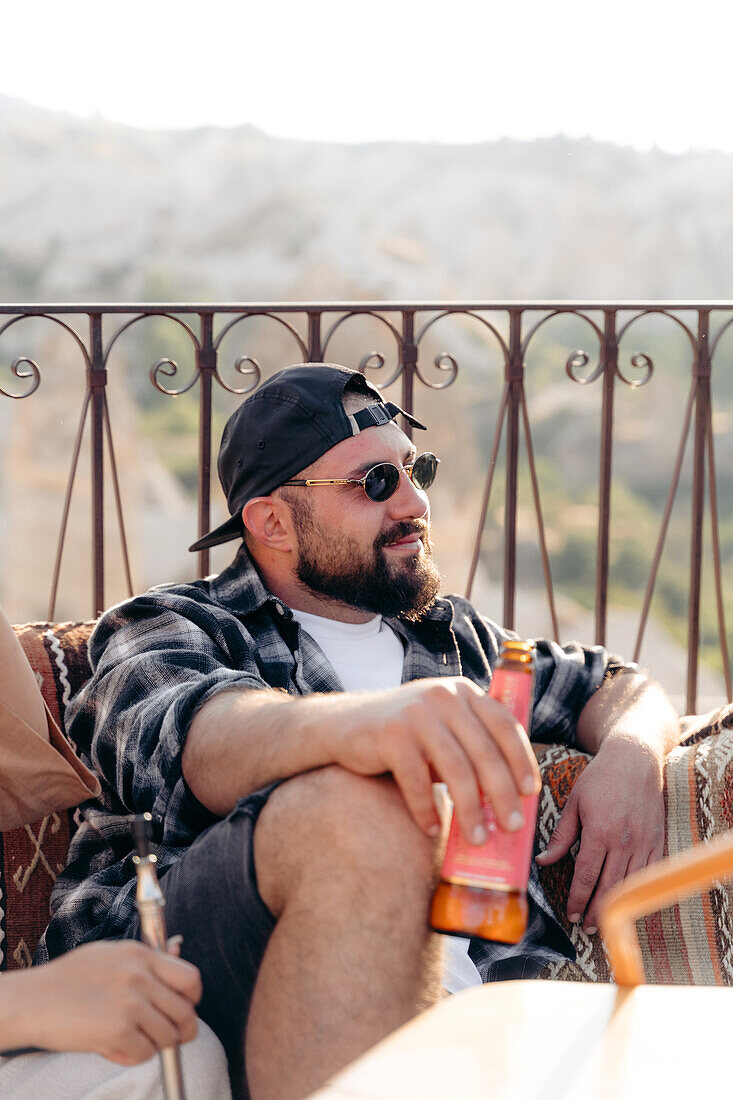 Erwachsener bärtiger Mann mit modischer Sonnenbrille sitzt mit einer Bierflasche neben einem Erntehelfer auf der Couch