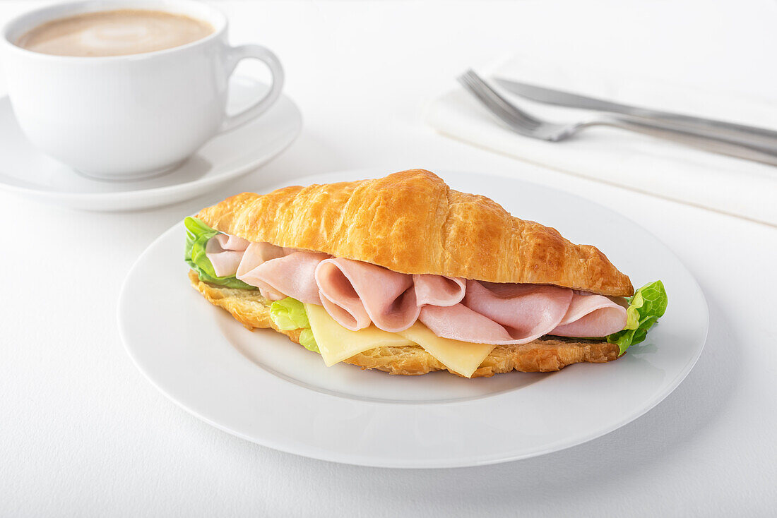 Leckeres Croissant mit Schinken und Käse auf weißem Teller neben einer Tasse Kaffee