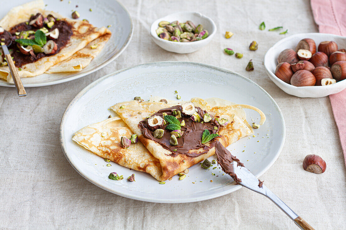 Draufsicht auf leckere Crêpes, die mit Schokolade und Nüssen garniert auf einem Teller auf dem Frühstückstisch serviert werden