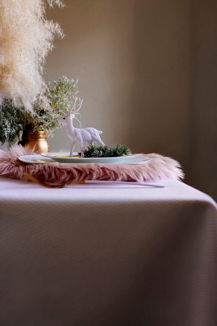 Von oben weihnachtlich gedeckter Tisch mit Kranzdekoration und weißem und goldenem Geschirr und weißem Rentier auf flauschigem Tischset auf rosa Tischtuch