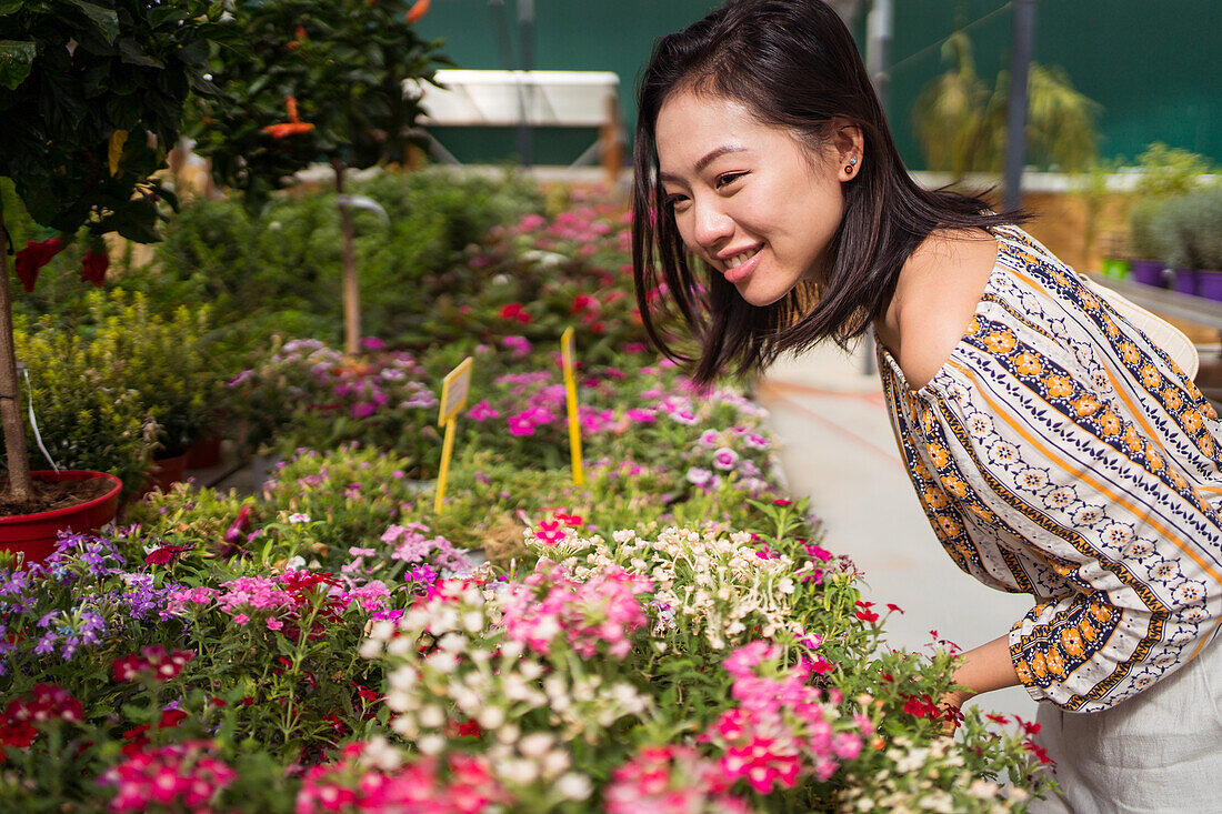 Seitenansicht einer fröhlichen jungen ethnischen Käuferin, die sich beim Pflücken blühender Blumen in einem Gartencenter nach vorne lehnt