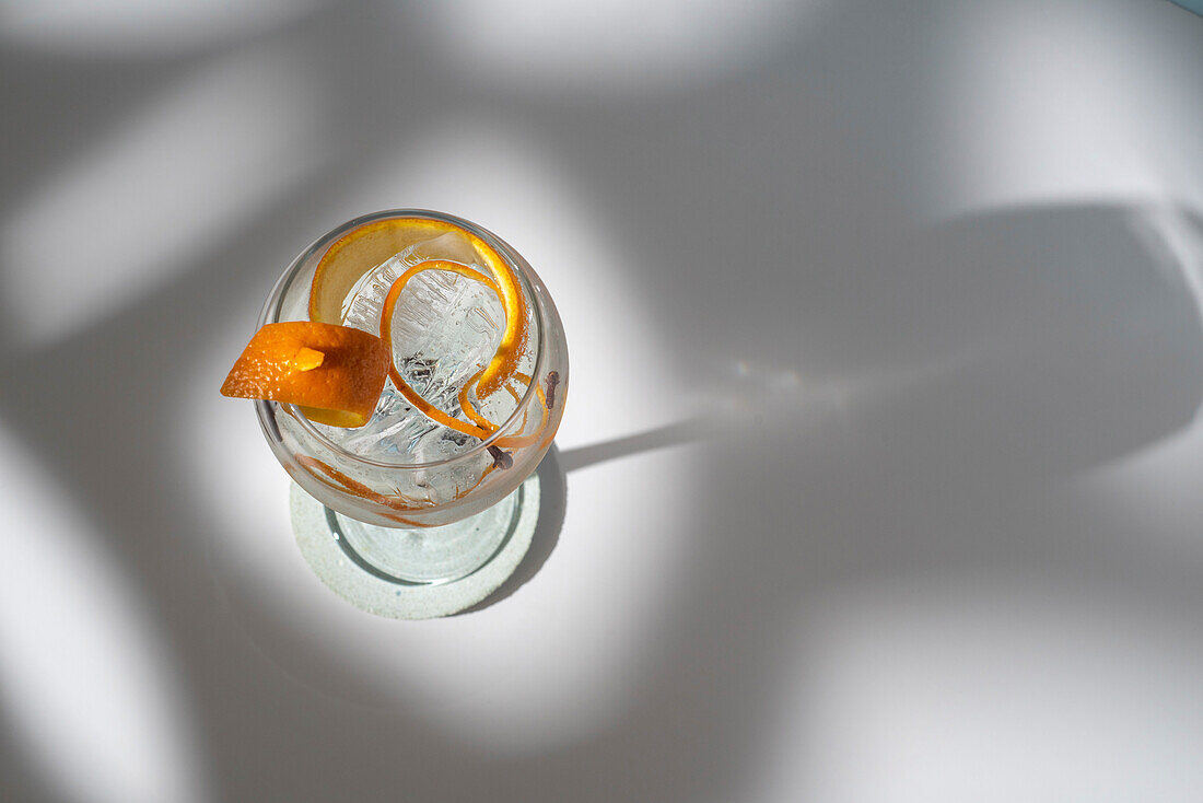 Draufsicht auf ein transparentes Glas mit einem Highball-Cocktail, dekoriert mit Zitrusfruchtschalen und Nelken im Schatten des Sonnenlichts