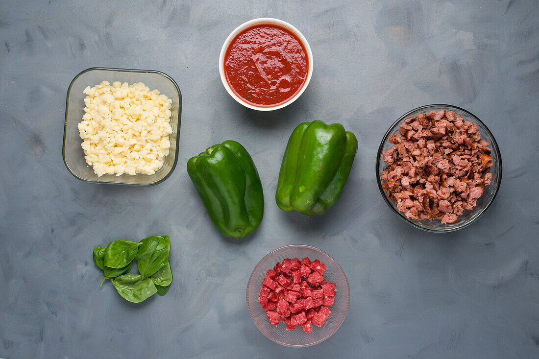 Top view of green bell peppers and ingredients for pizza stuffing with meat and shredded cheese on gray background in kitchen