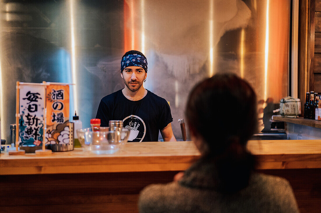 Rückenansicht einer gesichtslosen schwarzhaarigen Frau im Pullover, die am Tresen einer gemütlichen Ramen-Bar sitzt