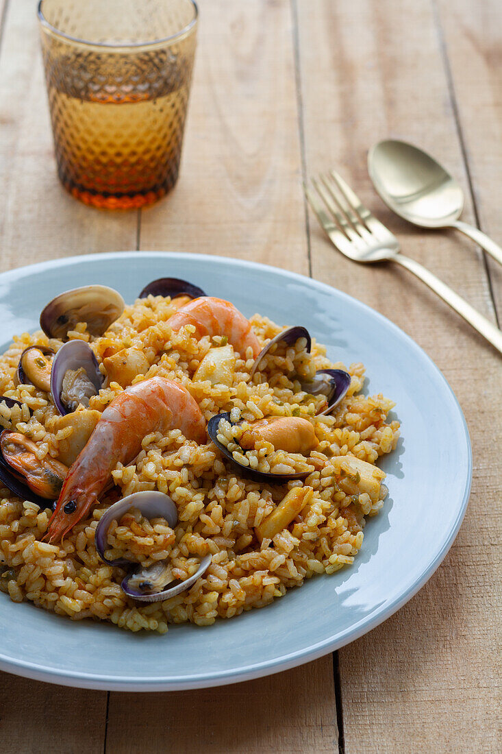 Plate with tasty fresh paella with mussels and shrimps served on wooden table with glass of refreshing beverage and cutlery