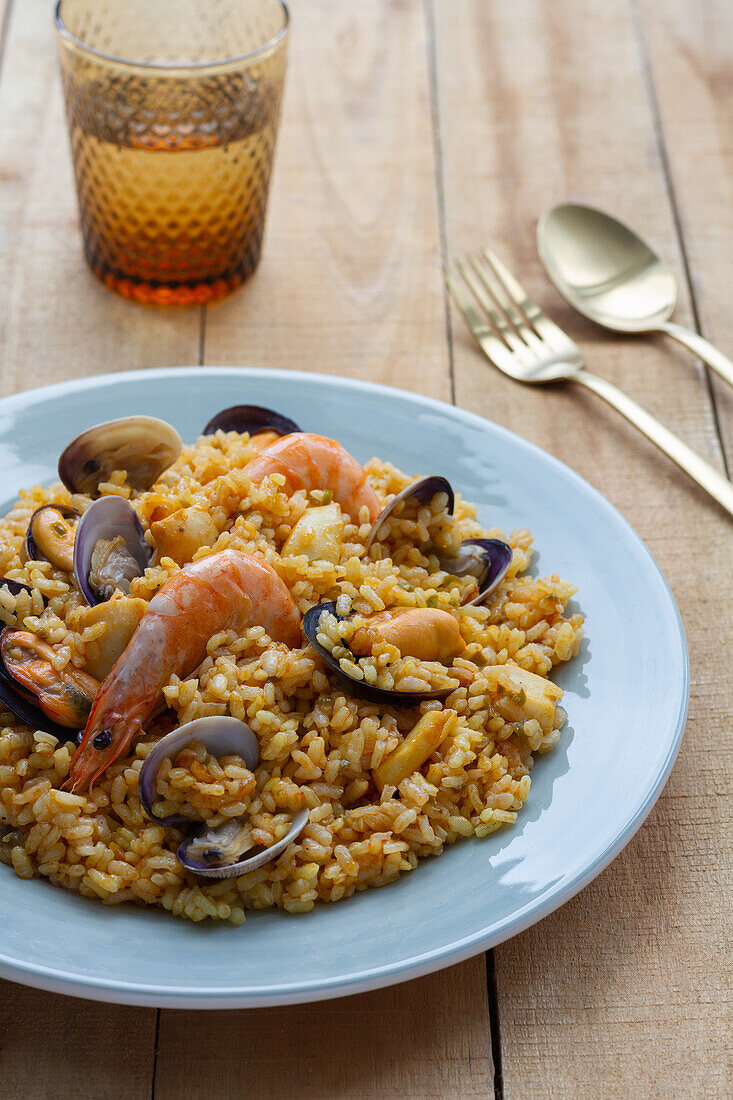 Teller mit leckerer frischer Paella mit Muscheln und Krabben, serviert auf einem Holztisch mit einem Glas eines erfrischenden Getränks und Besteck