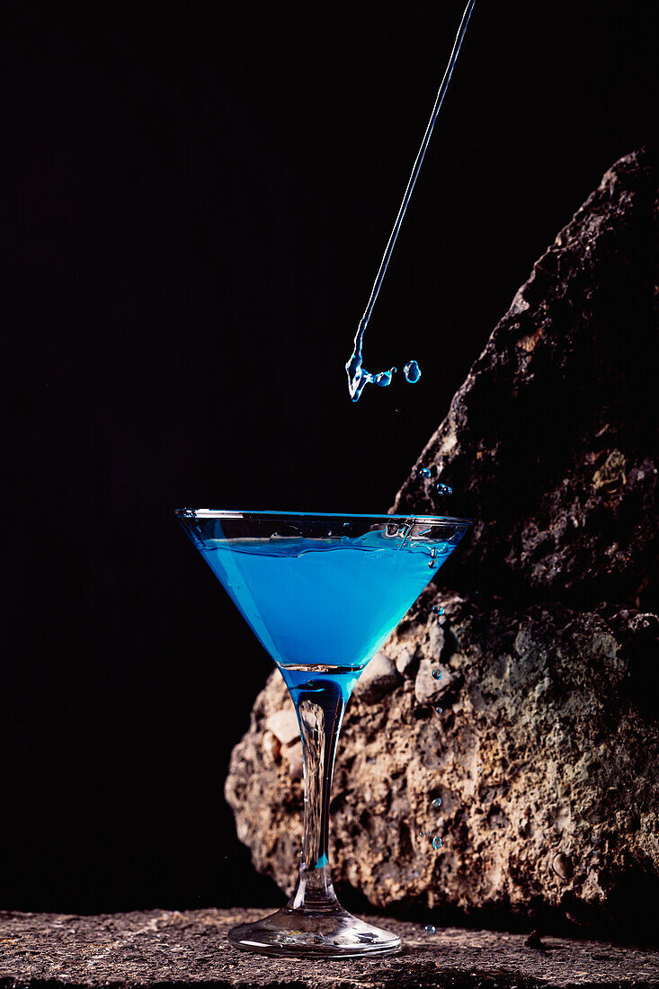 Tropfen eines blauen Lagunencocktails in einem eleganten Kristallglas auf einer rauen Oberfläche vor einem schwarzen Hintergrund