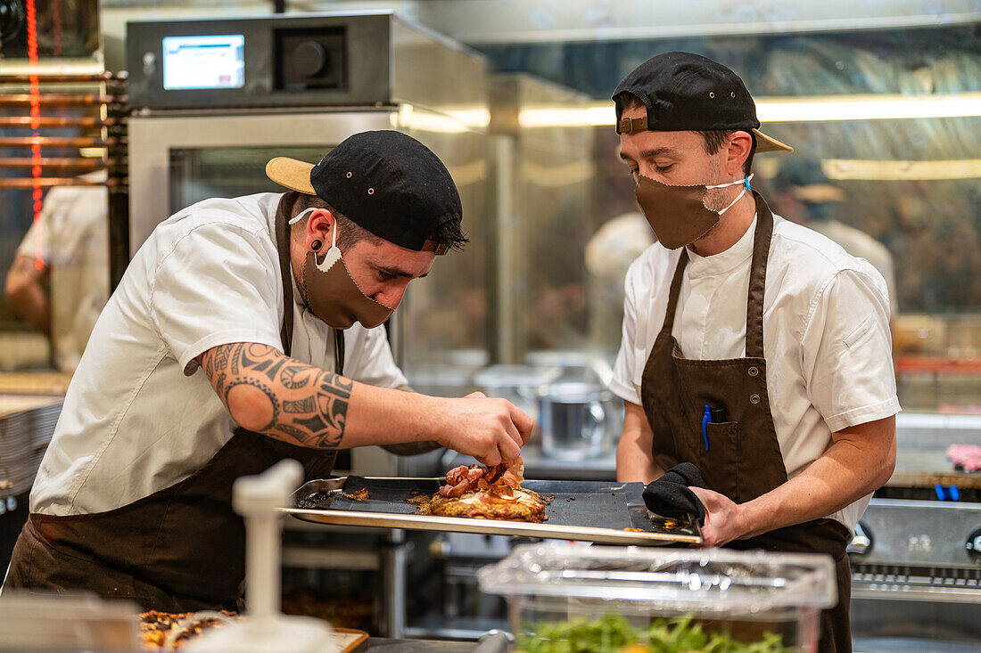 Männlicher Koch gibt Füllung auf Fladenbrot auf rostfreiem Tablett in die Hände eines Kollegen mit Schutzmaske und Uniform in einem Restaurant
