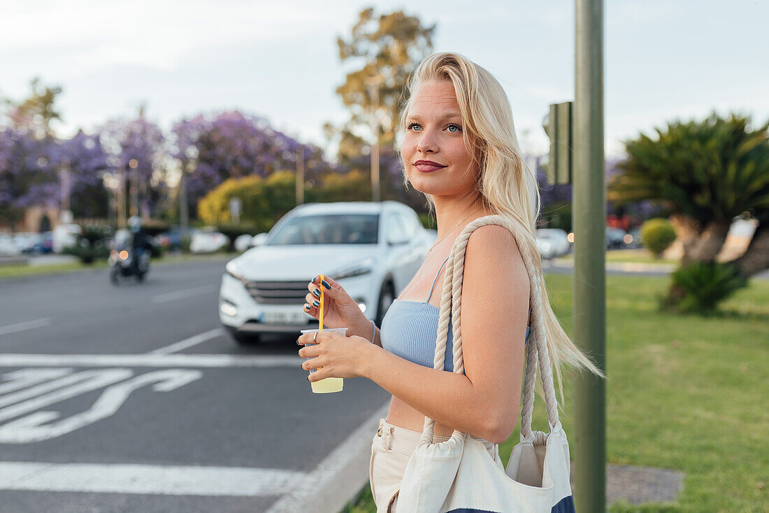 Side view of cheerful female standing with cold lemonade in plastic cup in street in summer