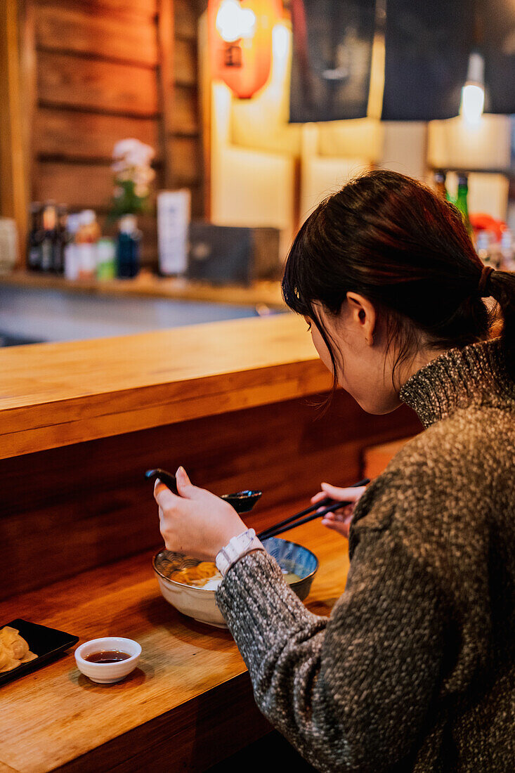 Side view of satisfied Asian woman in sweater smiling while taking spoon from worker sitting at wooden counter in ramen bar