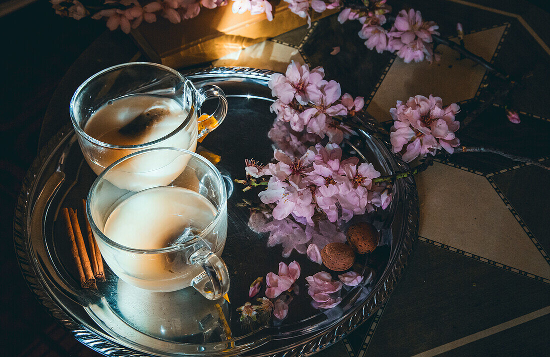 From above of glass cups full of tea with milk served on tray with cinnamon sticks and branches of flowers in room with sunbeam