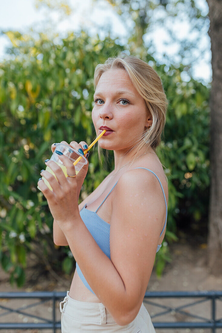 Seitenansicht einer charmanten Frau, die eine kalte, erfrischende Limonade mit Strohhalm trinkt, während sie im Sommergarten chillt und in die Kamera schaut