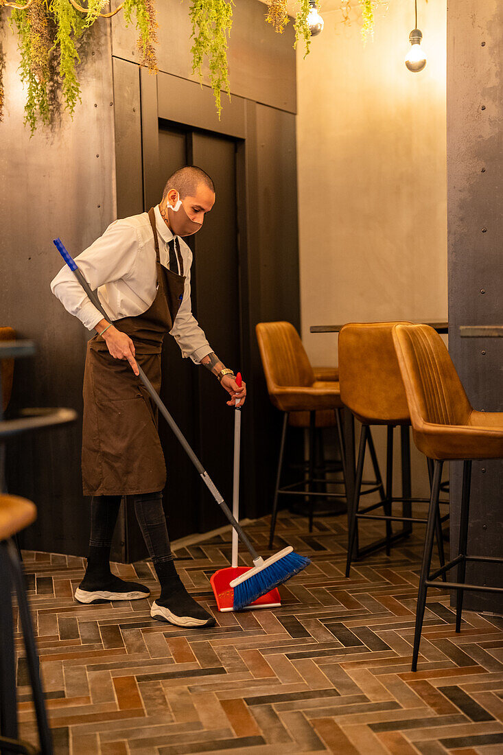 Beschäftigter männlicher Kellner mit Schaufel und Besen beim Reinigen des Fußbodens in einem modernen Restaurant während einer Coronavirus-Pandemie