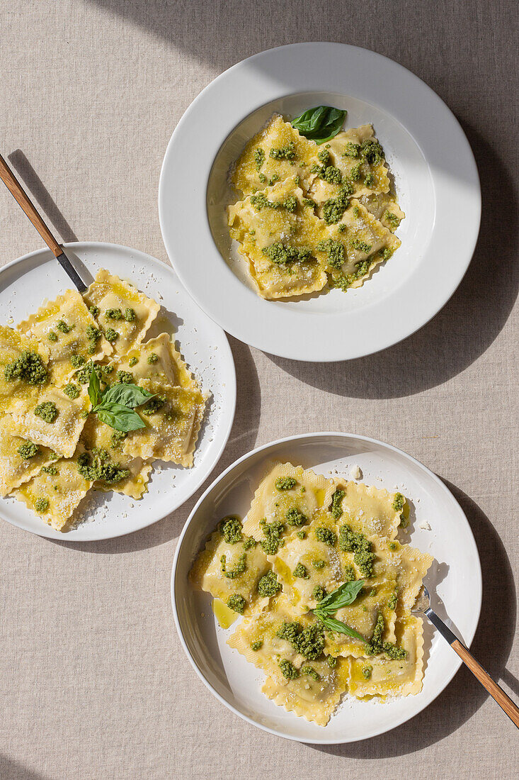 Draufsicht auf appetitlich gekochte Ravioli mit grüner Soße und Kräutern auf weißen Tellern mit Gabeln auf dem Tisch