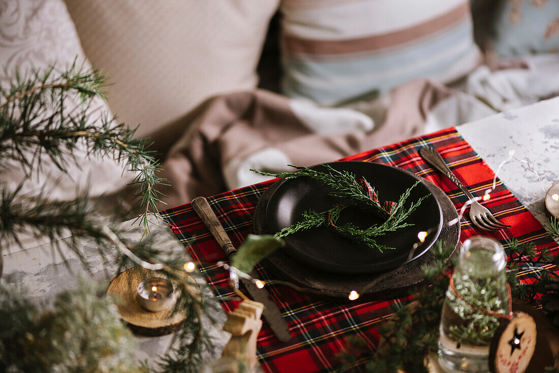 Weihnachtlich gedeckter Tisch mit Kranz auf Keramikteller auf rot kariertem Tischtuch im Hintergrund mit Lichtern