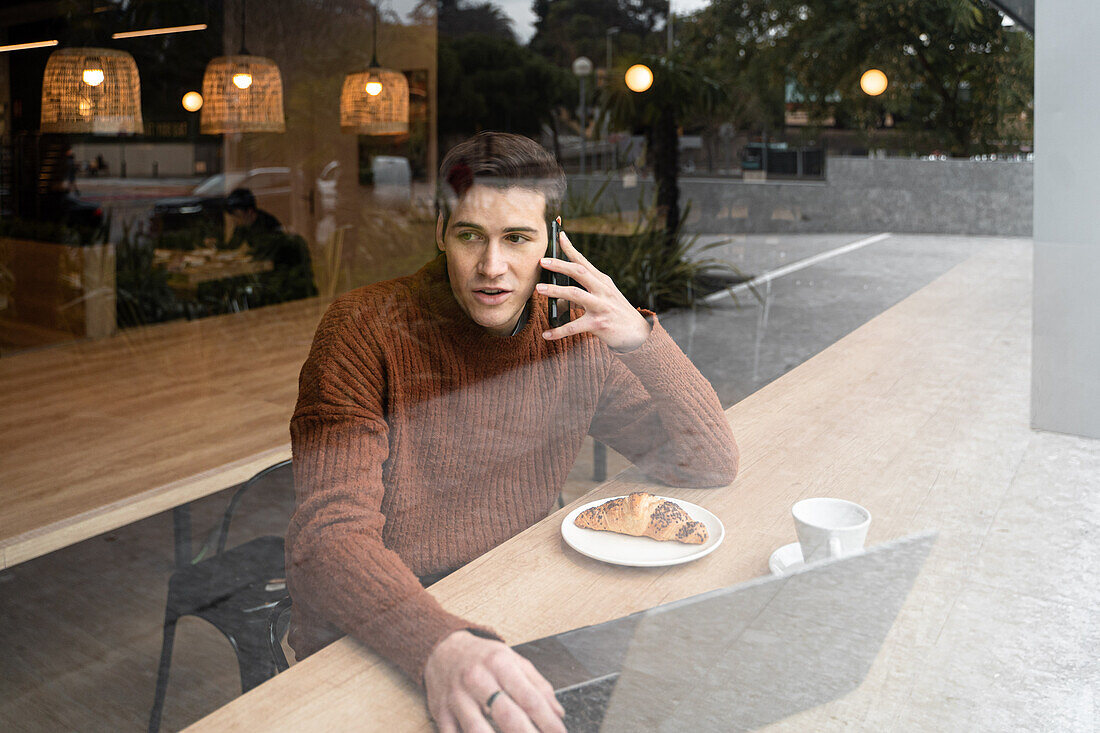 Blick durch das Fenster auf einen glücklichen jungen männlichen Studenten, der mit einem Laptop und einer Tasse Kaffee mit Croissant am Tisch sitzt und mit seinem Handy telefoniert, während er seine Zeit in einer modernen Cafeteria verbringt