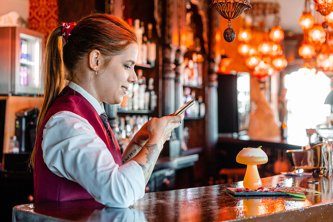 Seitenansicht einer fröhlichen Barkeeperin, die einen sauren Cocktail im Glas auf dem Tresen eines Pubs fotografiert