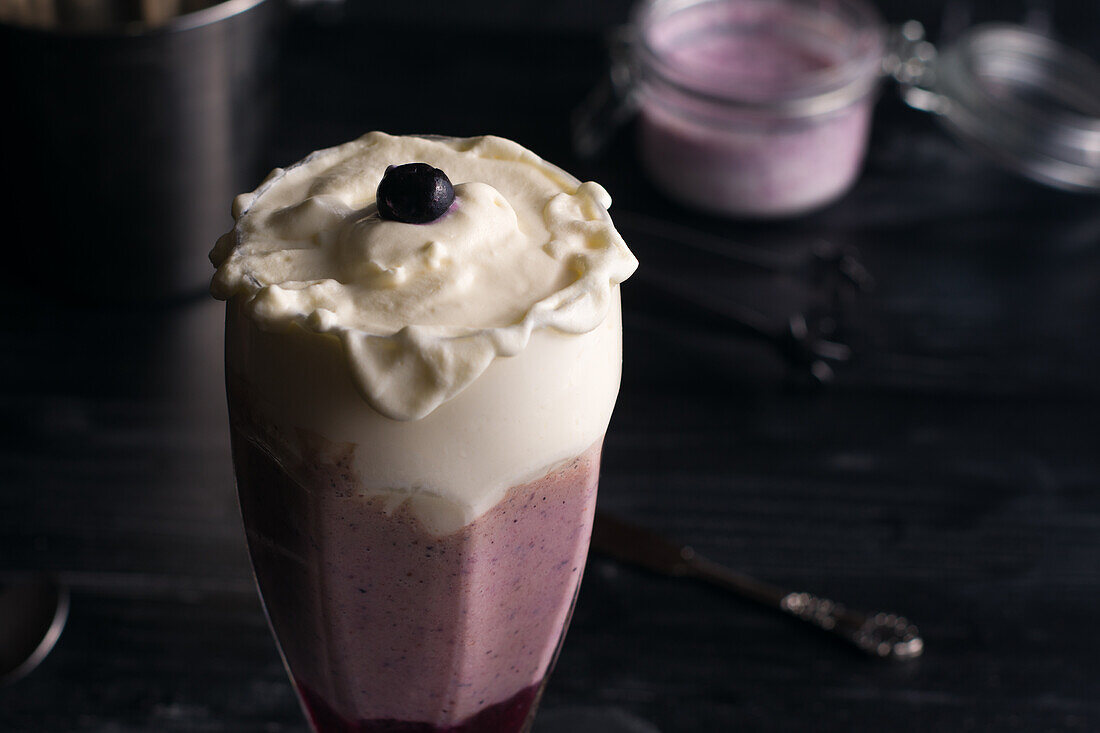 Glas köstlicher Bananen- und Blaubeer-Smoothie mit Schlagsahne auf Schneidebrett vor Joghurtglas und Metalleimer