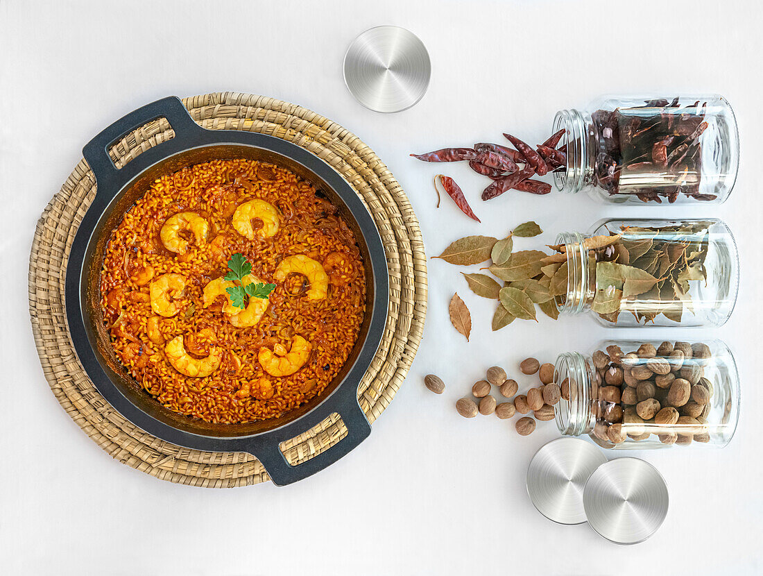 From above of tasty rice paella with shrimps in pot placed on wicker stand and glass bottles with red hot pepper bay leaves and nuts