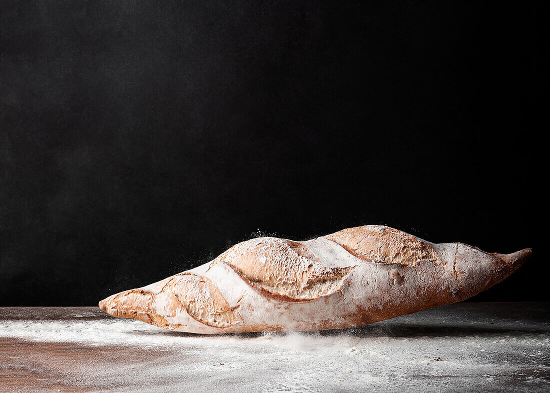 Appetitliches, frisch gebackenes Baguette mit knuspriger Kruste auf einem Holztisch mit weißem Mehl bedeckt vor schwarzem Hintergrund