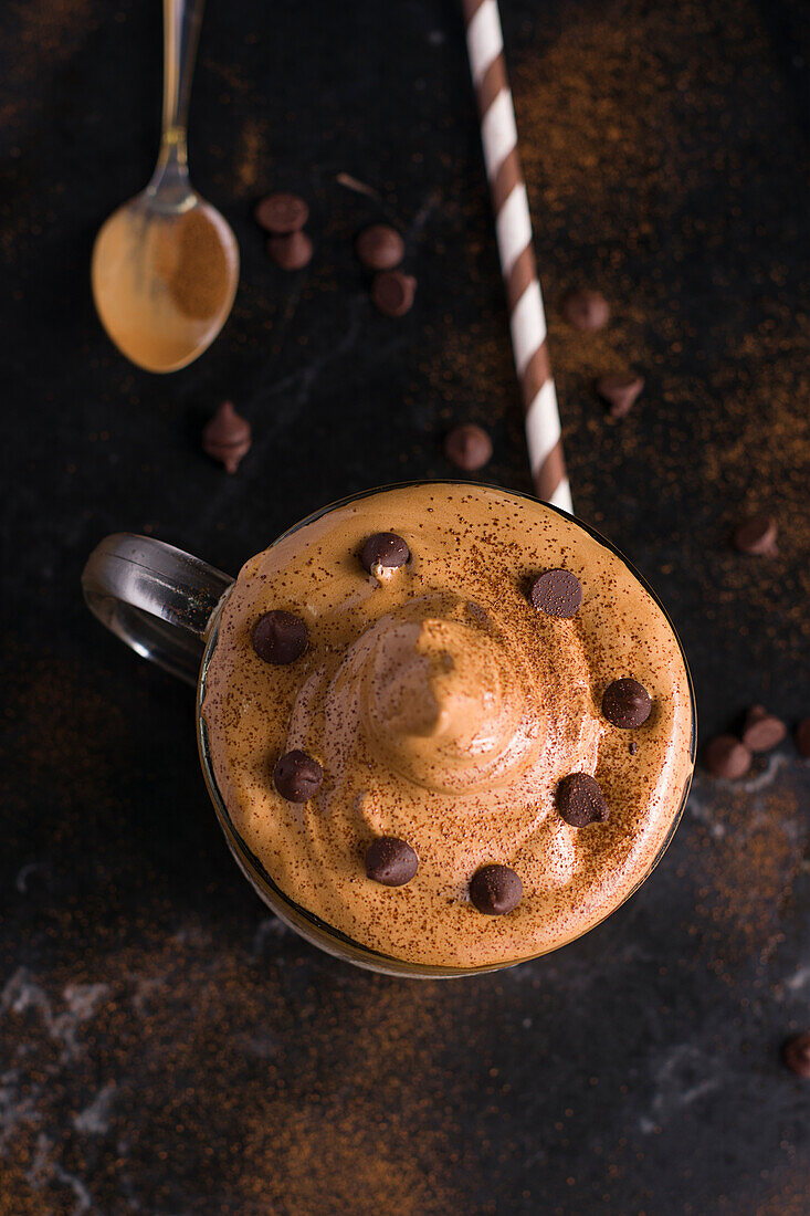 Von oben ein Glas süßer Dalgona-Kaffee mit schaumigem Belag, serviert auf einem Tisch mit Schokoladenwaffelrolle und Kakaopulver