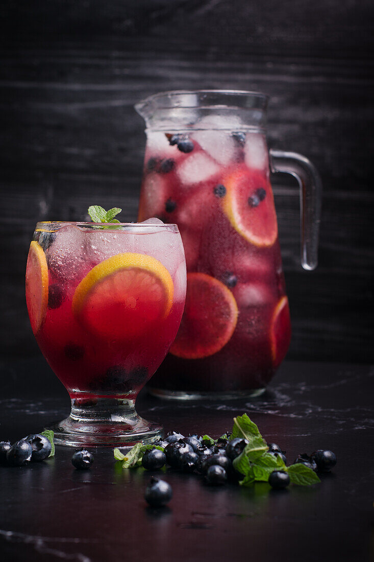 Glas und Krug mit erfrischender kalter Limonade mit frischen Blaubeeren und Zitronenscheiben auf einem dunklen Tisch