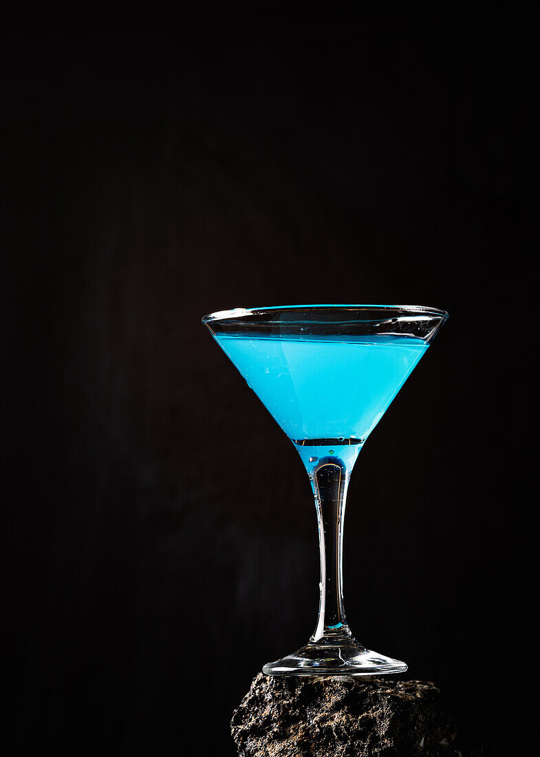 Blaue-Lagune-Cocktail in elegantem Kristallglas auf rauer Oberfläche vor schwarzem Hintergrund