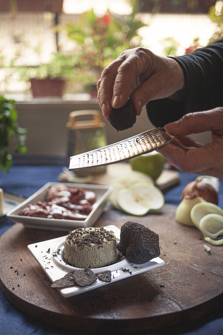 Unbekannte Köchin bestreut Weichkäse mit Trüffel mit einer Reibe, während sie am Tisch im Haus kocht