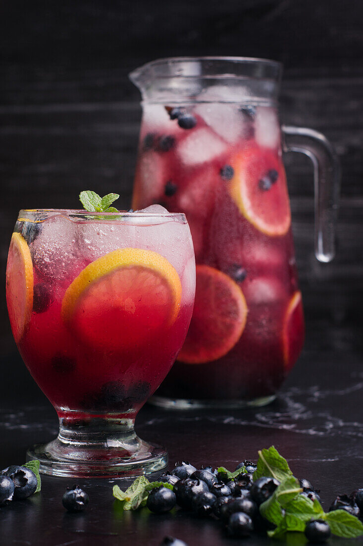 Glas und Krug mit erfrischender kalter Limonade mit frischen Blaubeeren und Zitronenscheiben auf einem dunklen Tisch