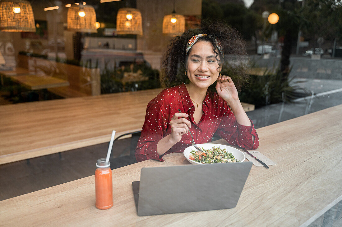 Through glass of positive young female blogger sitting at table with healthy meal and using laptop while spending time in modern restaurant