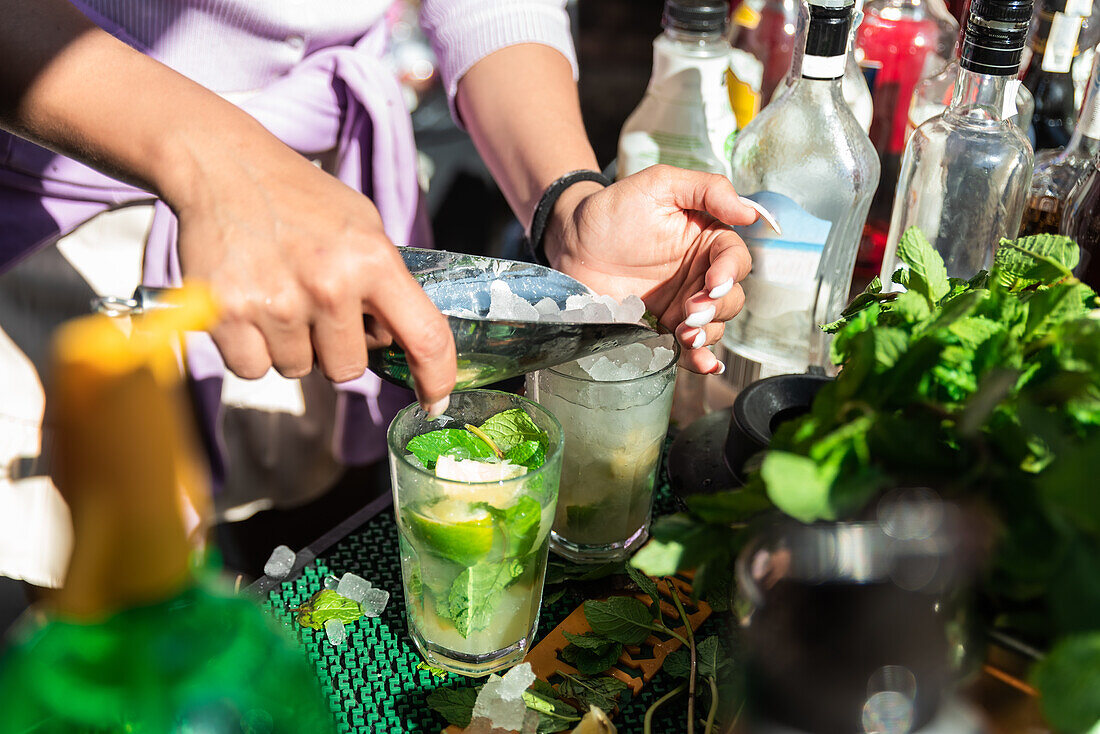 Crop anonymous female barkeeper adding ice cubes into glass while preparing cold refreshing mojito cocktails in sunny outdoor bar