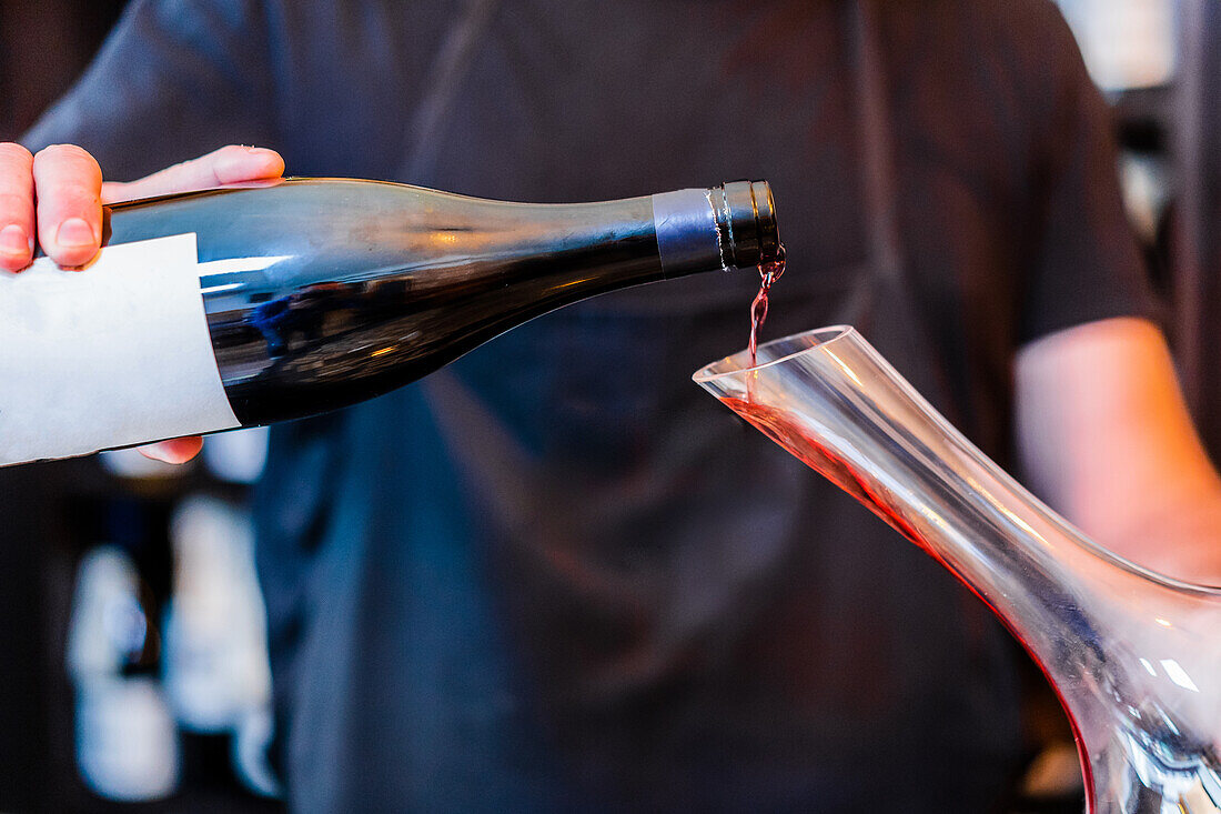 männlicher Sommelier in schwarzer Schürze, der am Bartresen steht und eine Flasche Rotwein in eine Glaskaraffe gießt, in einem Restaurant