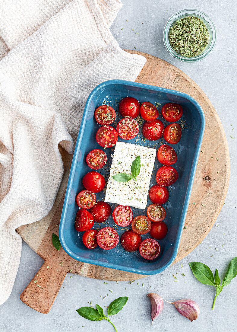 Reife Kirschtomaten mit schwarzen Olivenscheiben und Fetakäse in einer Auflaufform neben ungekochten Penne von oben