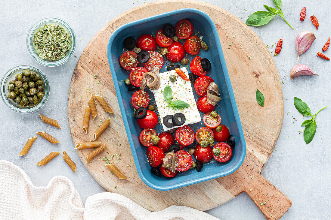 Reife Kirschtomaten mit schwarzen Olivenscheiben und Fetakäse in einer Auflaufform neben ungekochten Penne von oben