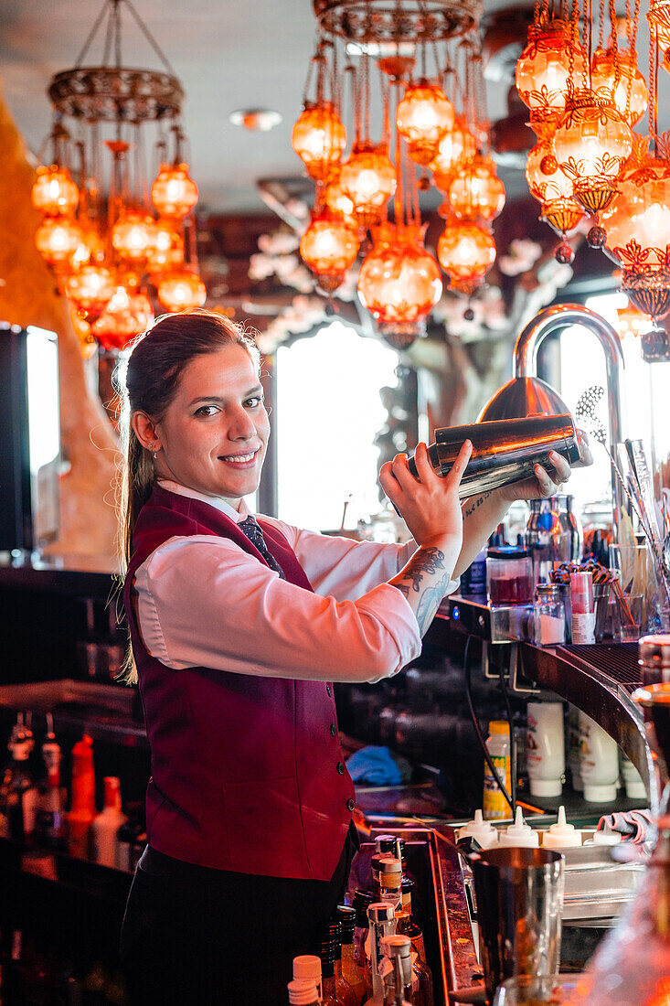 Seitenansicht einer Barkeeperin in Uniform, die einen Cocktail in einem Metallshaker schüttelt, während sie am Tresen eines Pubs steht und in die Kamera schaut