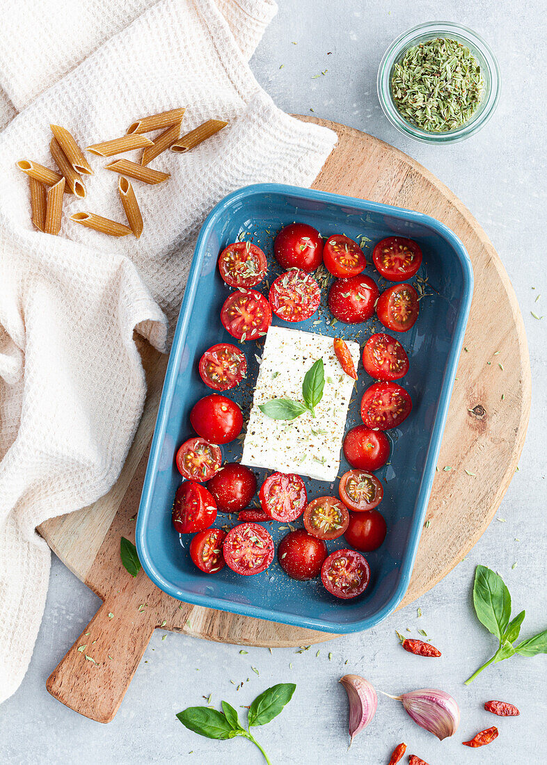 Reife Kirschtomaten mit schwarzen Olivenscheiben und Fetakäse in einer Auflaufform neben ungekochten Penne von oben