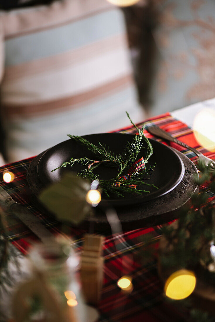 Weihnachtlich gedeckter Tisch mit Kranz auf Keramikteller auf rot kariertem Tischtuch im Hintergrund mit Lichtern