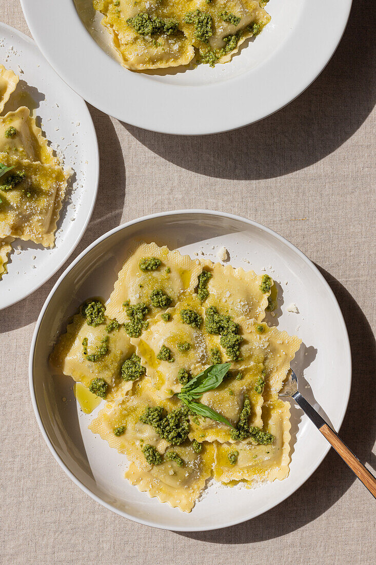 Draufsicht auf appetitlich gekochte Ravioli mit grüner Soße und Kräutern auf weißen Tellern mit Gabeln auf dem Tisch