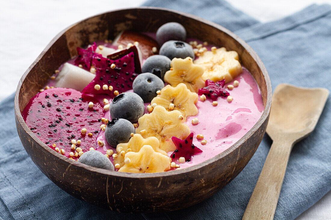 Blick von oben auf eine Schüssel mit einem leckeren Smoothie neben reifen Bananen- und Drachenfruchtscheiben mit Heidelbeeren zum Frühstück