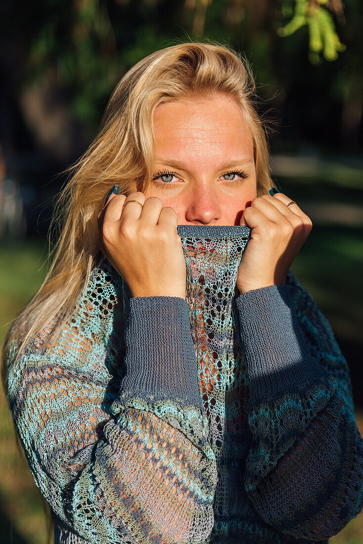 Zarte Frau mit blondem Haar und Strickpulli blickt im Park bei Sonnenuntergang in die Kamera