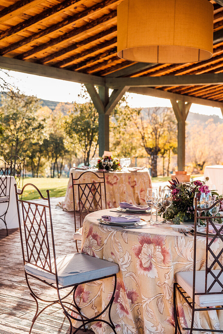 Geräumige Veranda mit Tellern, Weingläsern und Besteck auf mit frischen Blumen geschmückten Tischen für eine Hochzeitsfeier