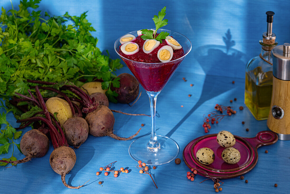 Glasbecher mit leckerer Rote-Bete-Creme, garniert mit Wachteleiern und Petersilie, neben Serviette mit Löffel und frischen Zutaten vor blauem Hintergrund