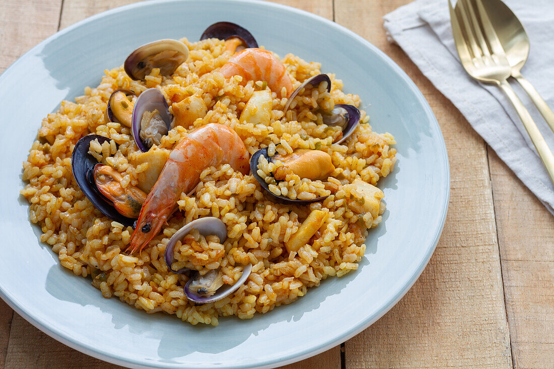 Teller mit leckerer frischer Paella mit Muscheln und Krabben, serviert auf einem Holztisch mit einem Glas eines erfrischenden Getränks und Besteck