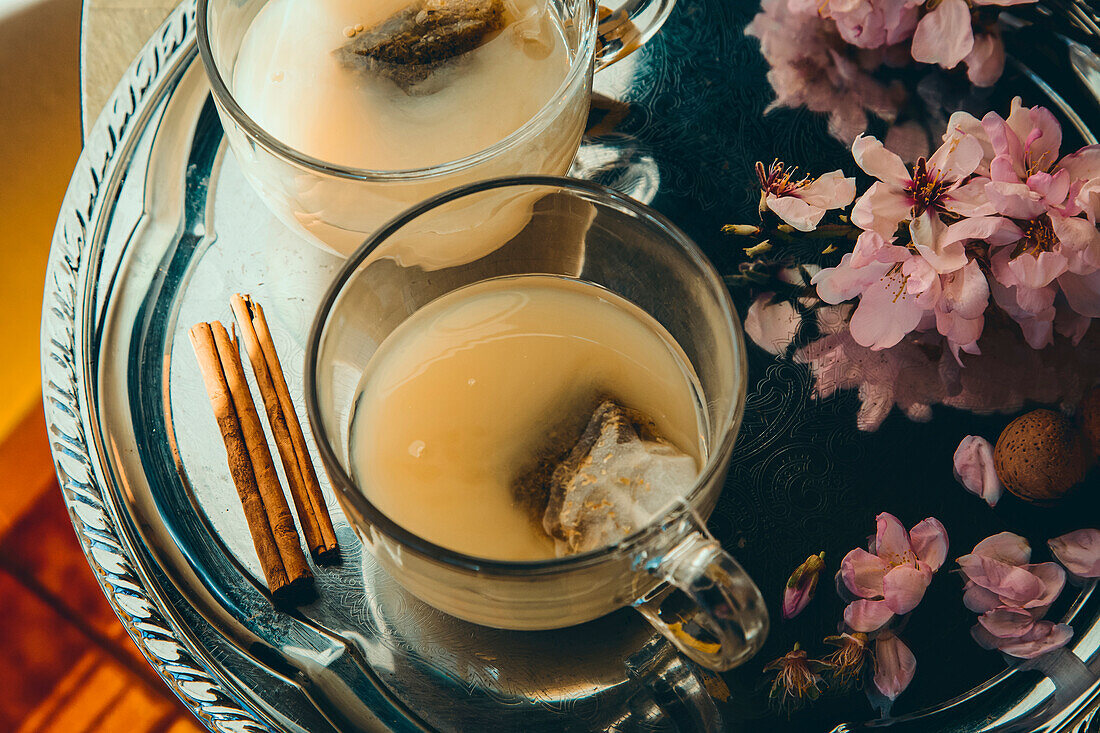 From above of glass cups full of tea with milk served on tray with cinnamon sticks and branches of flowers in room with sunbeam