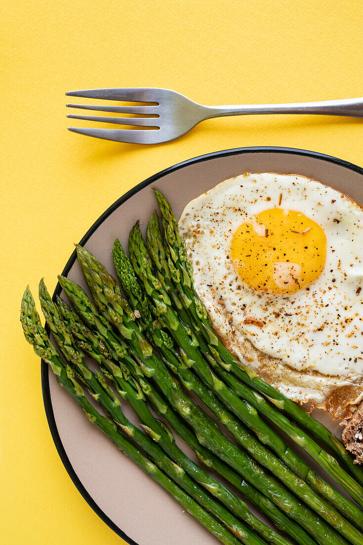From above breakfast dish of fried egg with green asparagus server on plate on yellow background