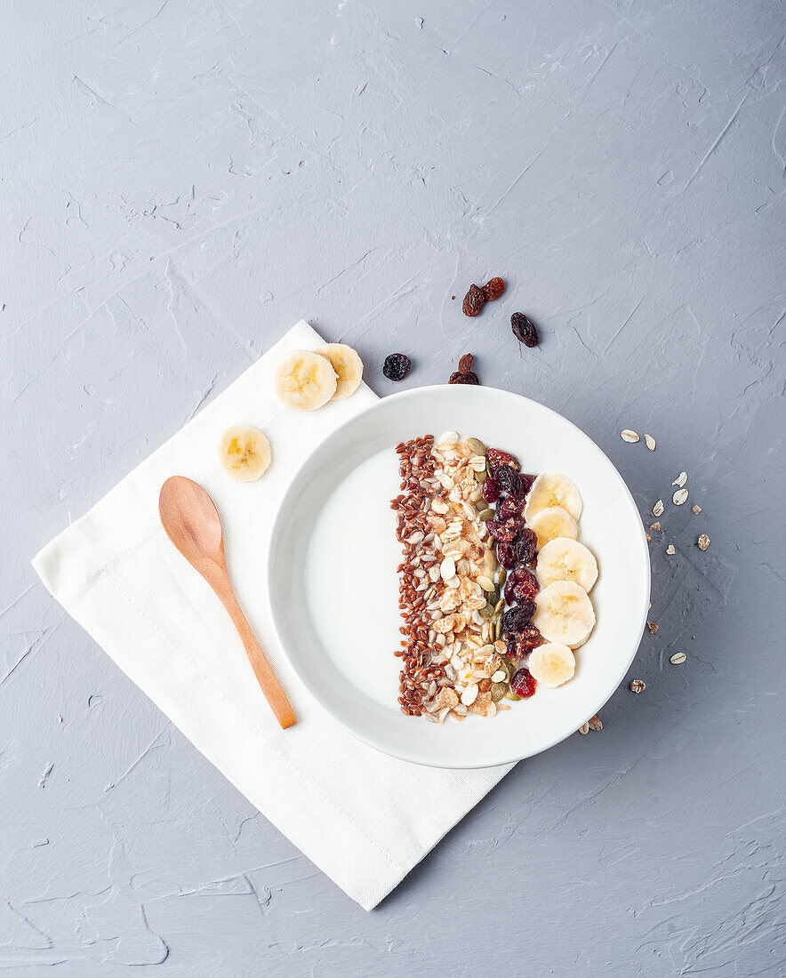 Draufsicht auf eine Schale mit weißem Joghurt mit verschiedenen Cerealien und geschnittener Banane, die zum gesunden Frühstück auf einem Holztisch mit Holzlöffel serviert wird