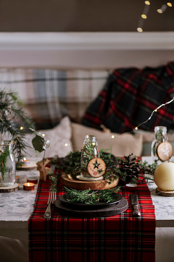 Weihnachtlich gedeckter Tisch mit Kranz auf dem Teller, dekorativem Holzschmuck und rot kariertem Tischtuch mit gelben Lichtern im Hintergrund