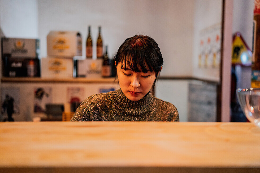 Young Asian woman in casual wear sitting at wooden counter while waiting for order in ramen bar