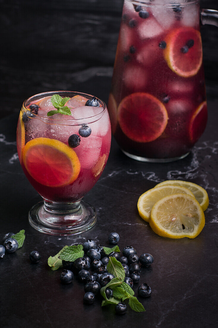 Hoher Winkel von Glas und Krug mit erfrischender kalter Limonade mit frischen Blaubeeren und Zitronenscheiben auf einem dunklen Tisch